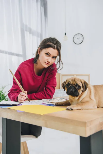 Junge Frau Zeichnet Während Mops Auf Ihrem Tisch Wartet — Stockfoto