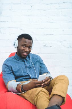 Afro-Amerikan yakışıklı ile müzik kulaklık ve fasulye çanta içinde oturan smartphone