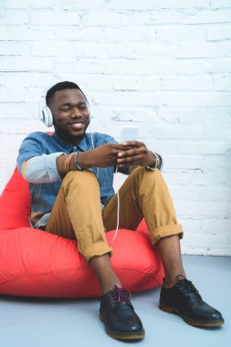 Afro-Amerikan man ise kulaklık müzik ve fasulye çanta içinde oturan Smartphone vasıl arıyorum