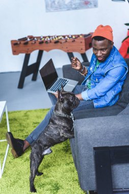 French bulldog by African american man sitting on sofa with laptop clipart