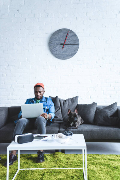 Digital devices on table in front of young man working on laptop and sitting om sofa by French bulldog