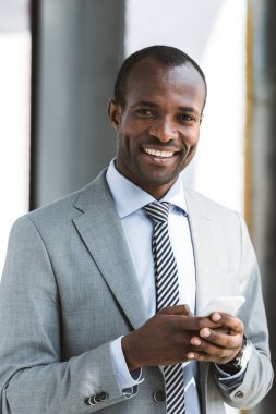 smartphone kullanarak ve kameraya gülümseyen yakışıklı genç Afro-Amerikalı işadamı portresi