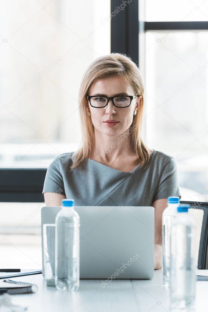 beautiful businesswoman in eyeglasses using laptop and looking at camera
