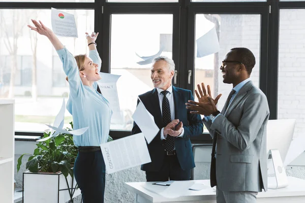 excited multiethnic business people applauding and throwing papers in office