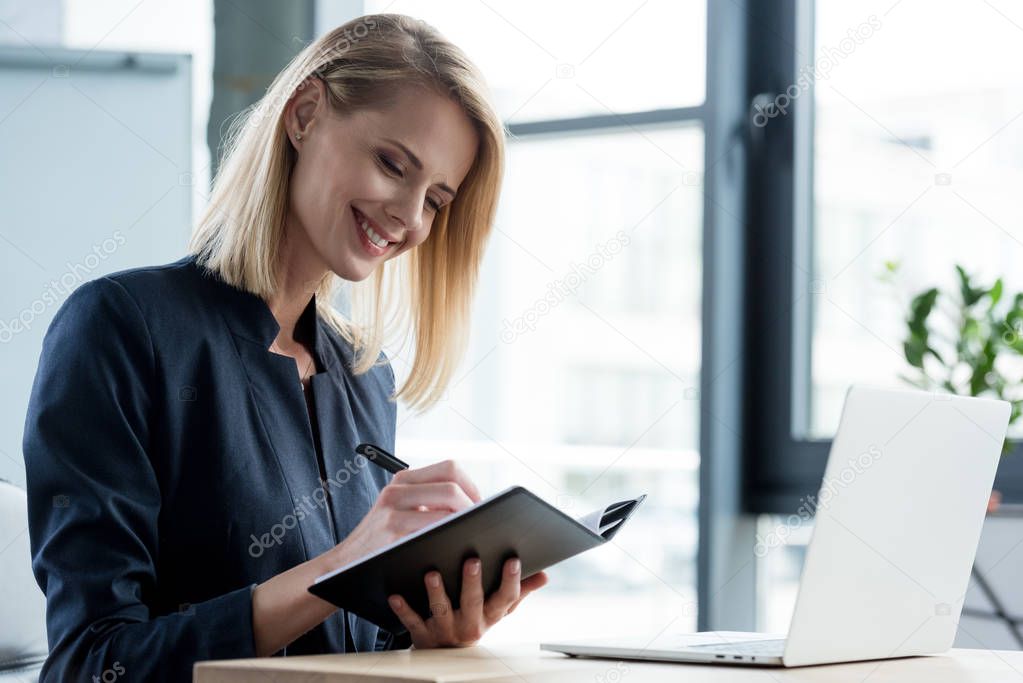 beautiful smiling businesswoman taking notes in notepad at workplace