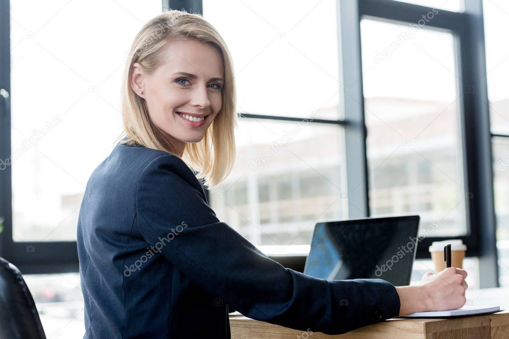 beautiful businesswoman smiling at camera while using laptop and taking notes in office
