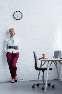 elegant senior businesswoman posing near workplace with laptop and coffee clipart