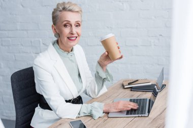 smiling senior businesswoman with disposable cup of coffee using laptop in office clipart