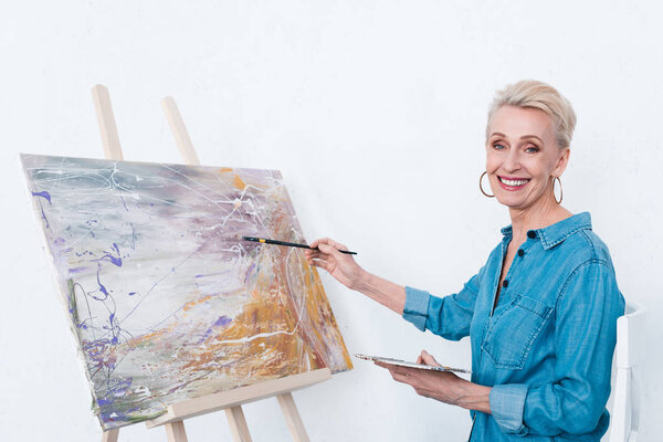 пожилая веселая женщина живопись с палитрой на мольберте в мастерской
