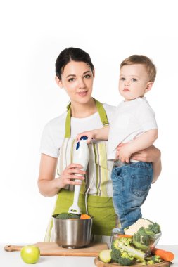 El blender ve beyaz arka plan üzerinde izole malzemelerle masada duran oğlu tutan anne 