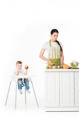 baby boy in highchair with toy and mother cutting zucchini at table isolated on white background  clipart