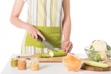 cropped shot of woman cutting zucchini by knife at table with puree jars, cauliflower and pumpkin  clipart