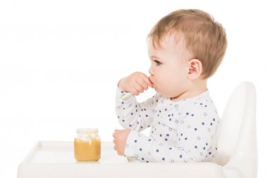  erkek bebek sandalyesi beyaz arka plan üzerinde izole oturuyor ve püre kavanozdan yeme yan görünüm 