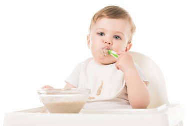 little boy in bib eating porridge by spoon and sitting in highchair isolated on white background  clipart