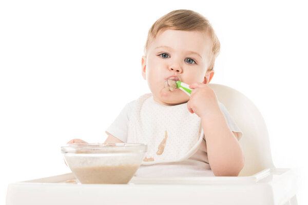 little boy in bib eating porridge by spoon and sitting in highchair isolated on white background 