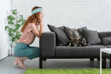 smiling young woman looking at her tabby cat while he sitting on couch at home clipart