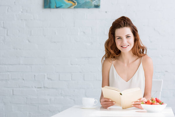 happy young woman sitting at table with coffee and strawberry and reading book at home