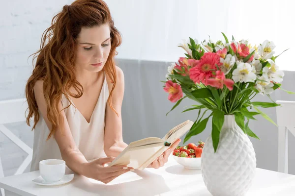 Concentrated young woman reading book while sitting at table with coffee cup and flowers in vase — Stock Image Concentrated Young Woman Reading Book While Sitting Table Coffee Cup Stock Picture