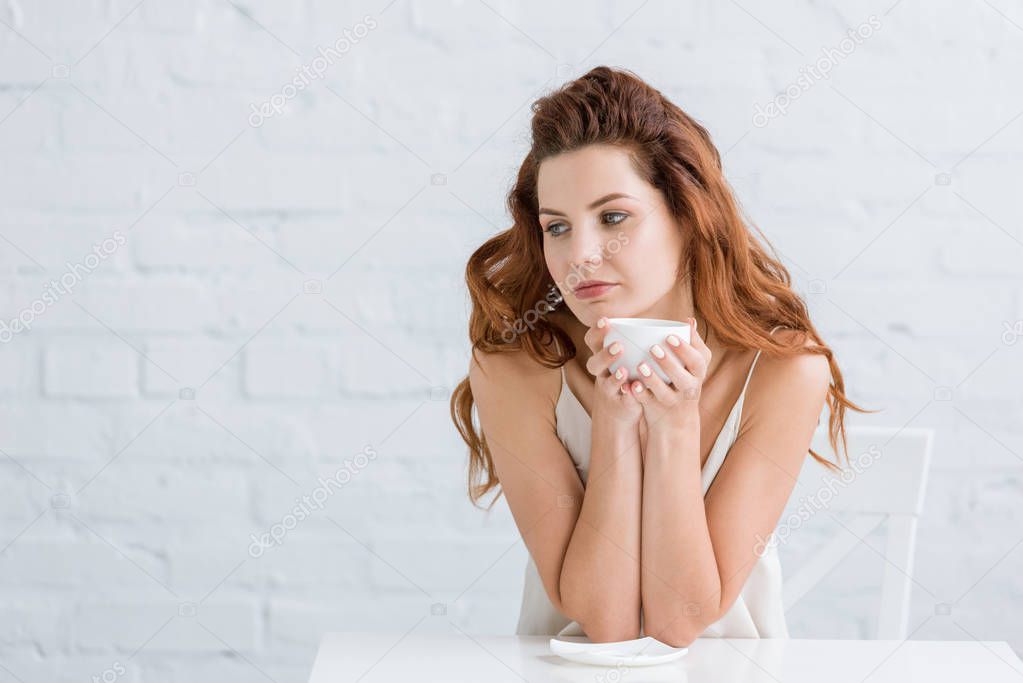 thoughtful young woman with cup of coffee in front of white brick wall