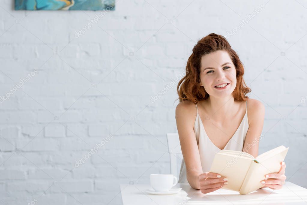 smiling young woman reading book with coffee cup on table at home