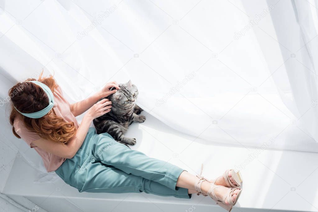 high angle view of attractive young woman petting adorable tabby cat while sitting on windowsill