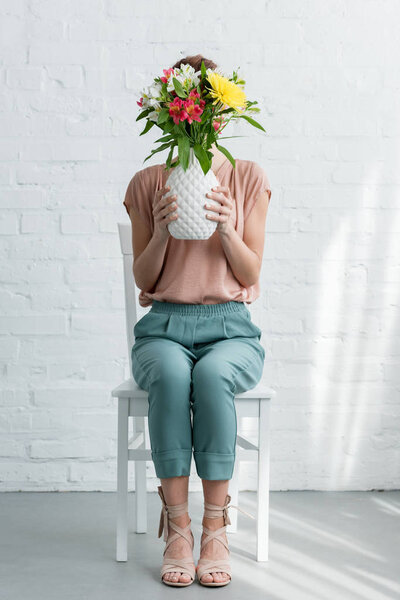 woman covering face with flowers in vase while sitting on chair in front of white brick wall
