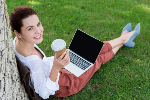 高角度视图的快乐年轻女子坐在草地上 用纸杯咖啡 看着相机 工作与笔记本电脑 — 图库照片