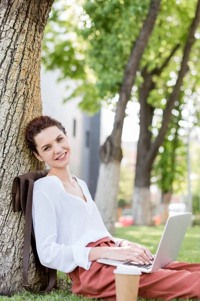 快乐的年轻自由职业者工作与笔记本电脑 而靠在树干在公园和看相机 — 图库照片