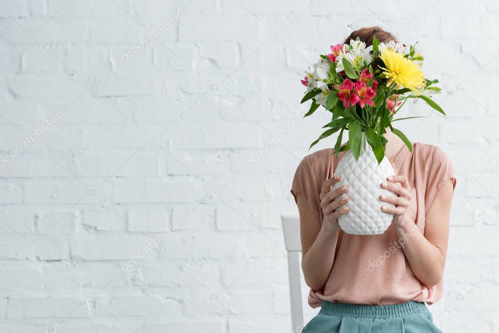 woman covering face with flowers in vase in front of white brick wall