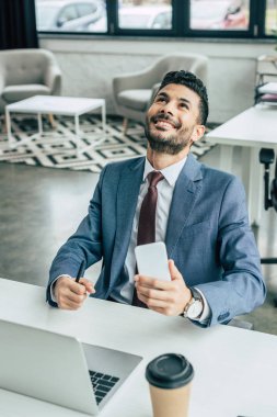 Mutlu iş adamı dizüstü bilgisayarın yanında oturmuş akıllı telefon tutarken yukarı bakıyor.