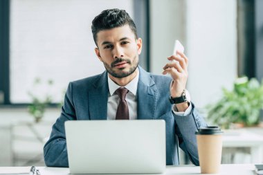 Sıkıcı iş adamı dizüstü bilgisayarın yanında oturmuş kameraya bakıyor ve elinde akıllı telefon tutuyor.