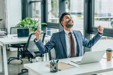 happy businessman looking up and showing winner gesture while sitting at workplace clipart