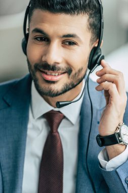 handsome call center operator touching headset while working in office clipart