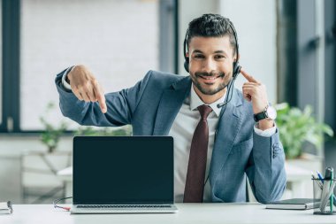 cheerful call center operator pointing with finger at laptop with blank screen  clipart