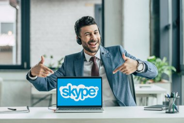 Kyiv, Ukrayna - 10 Ekim 2019: Neşeli çağrı merkezi operatörü kameraya göz kırpıyor ve ekranda Skype olan bilgisayarı işaret ediyor.
