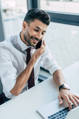 Gülümseyen iş adamı akıllı telefondan konuşuyor ve dizüstü bilgisayar kullanıyor.