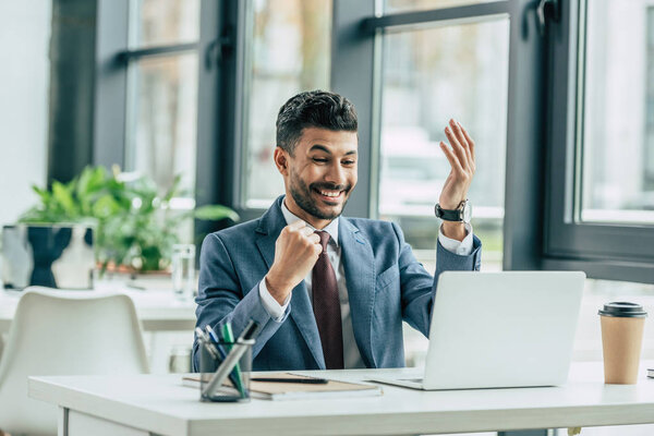 excited businessman showing winner gesture while sitting at workplace near laptop