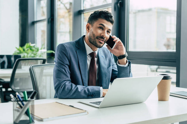 smiling businessman talking on smartphone while sitting at workplace near laptop