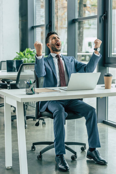 happy businessman screaming and showing winner gesture while sitting at workplace