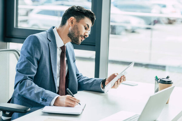 concentrated businessman looking at digital tablet and writing in notebook