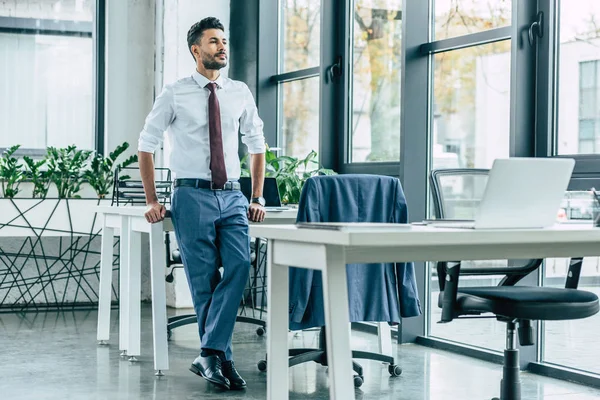 young confident businessman standing near desk and looking away