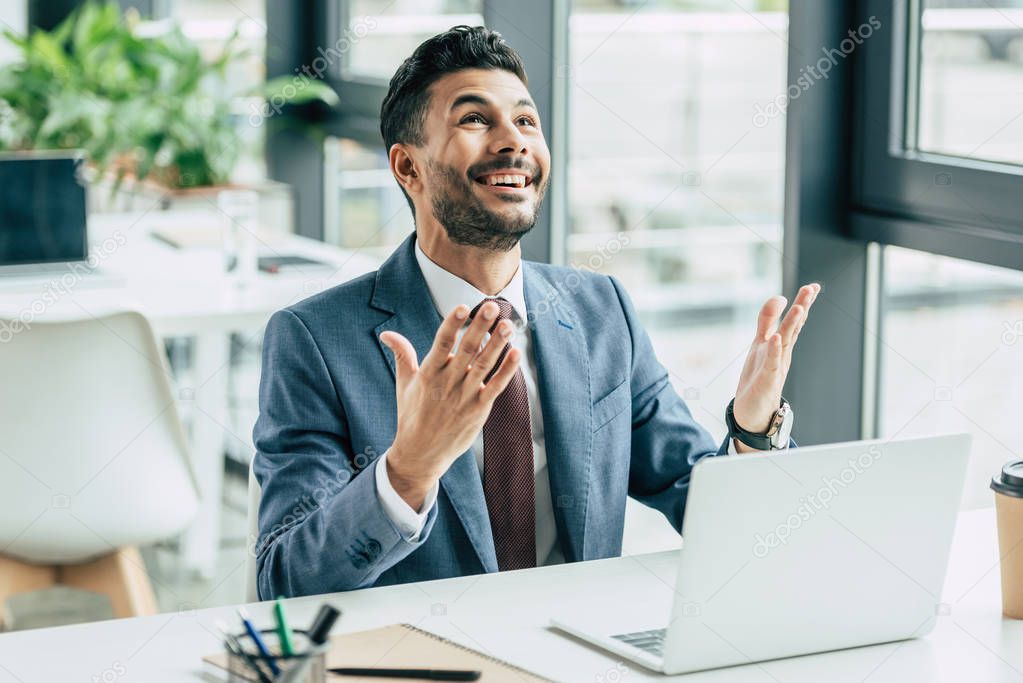 smiling businessman showing please gesture and looking up while sitting at workplace