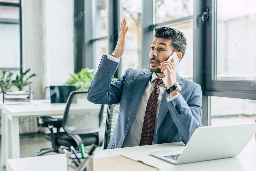 discouraged businessman showing indignation gesture while sitting at workplace and talking on smartphone