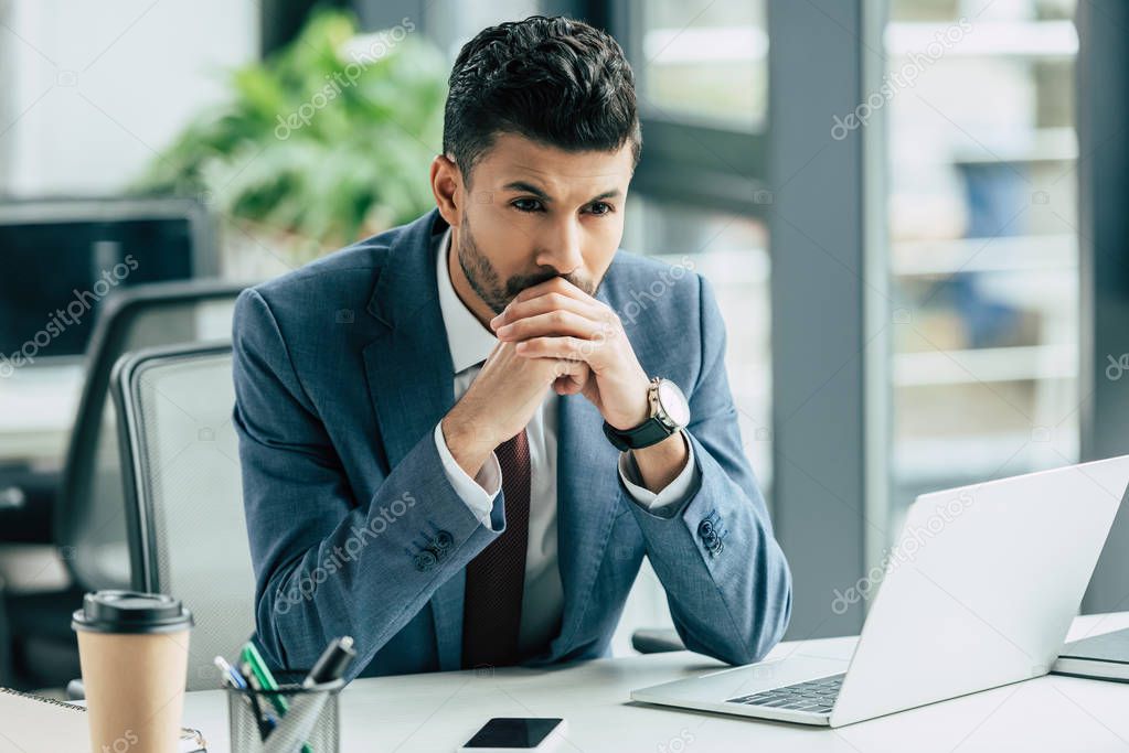 pensive businessman sitting at workplace near laptop and looking away