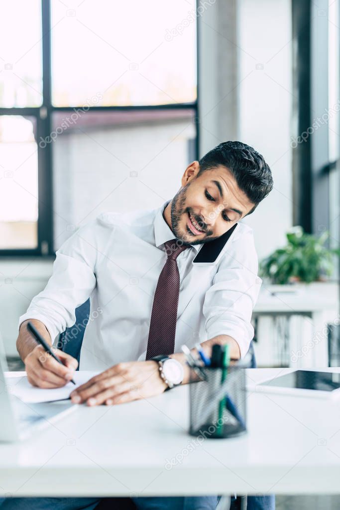 selective focus of cheerful businessman talking on smartphone and using laptop