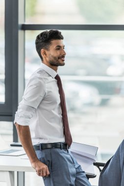 Genç işadamı işyerinde dururken gülümsüyor ve başka tarafa bakıyor.