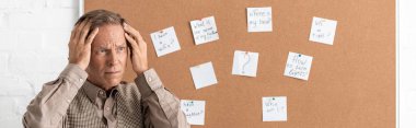 panoramic shot of retired man with alzheimer disease touching head and standing near board with papers and letters  clipart