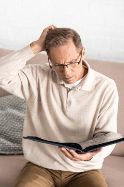 thoughtful man in glasses with alzheimer disease holding photo album at home  clipart