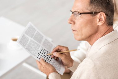 selective focus of senior man in glasses holding pencil near crossword in newspaper  clipart