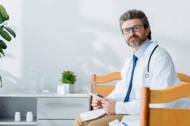 Beyaz önlüklü yakışıklı doktor yatakta oturuyor ve hastanede akıllı telefon tutuyor. 